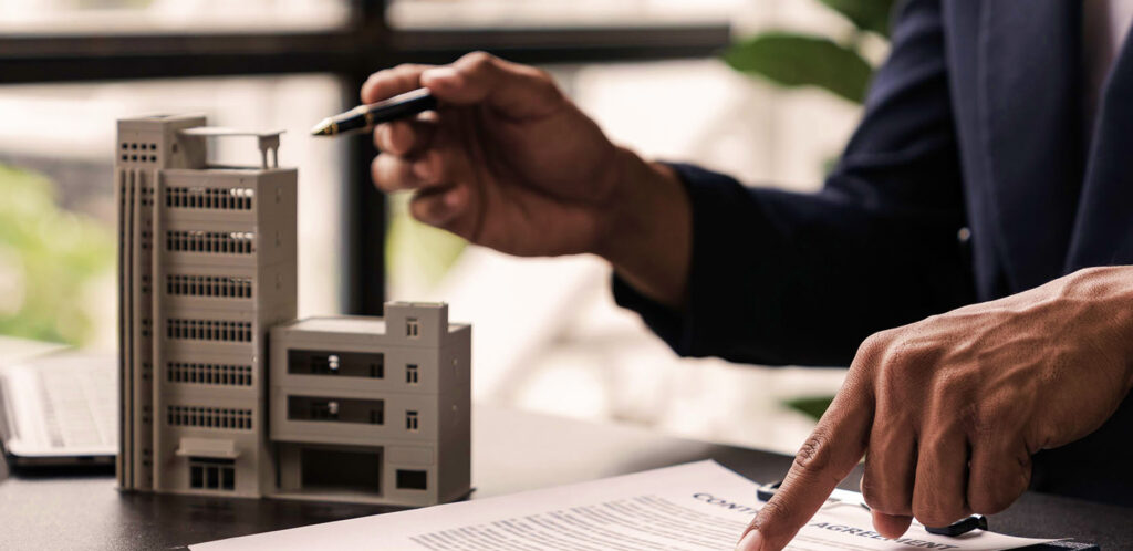 Eine Person im Anzug hält einen Stift in der Hand und zeigt auf Vertragsunterlagen. Auf dem Tisch steht ein Modellhaus.