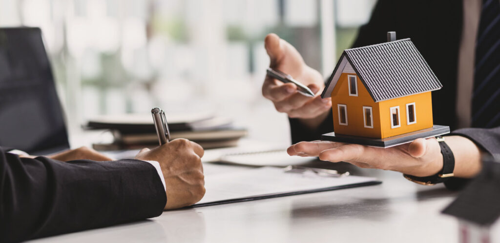 Ein Mann im Anzug hält ein Modellhaus in der Hand. Gegenüber sitzt eine Person und unterzeichnet eine Vertrag.